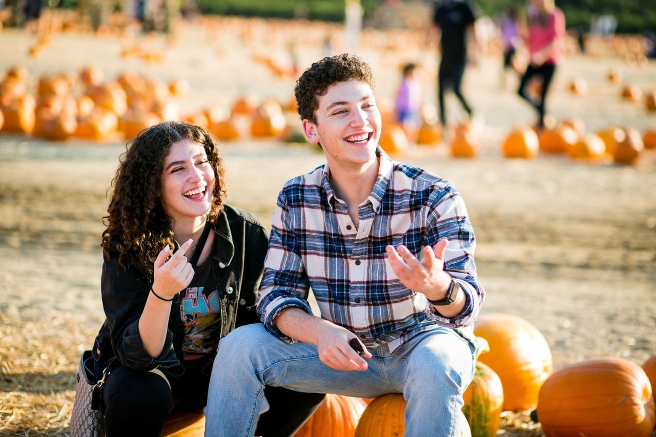 Two visitors sitting on pumpkins and laughing together in a pumpkin patch.
